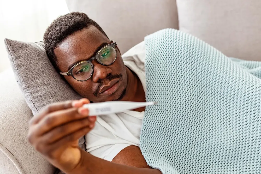 Man lying on the sofa under a blanket reading a thermometer